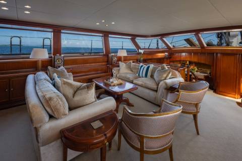 Plush sofas and armchairs fill the foreground of a luxurious yacht salon, while large windows in the background reveal a serene ocean view.