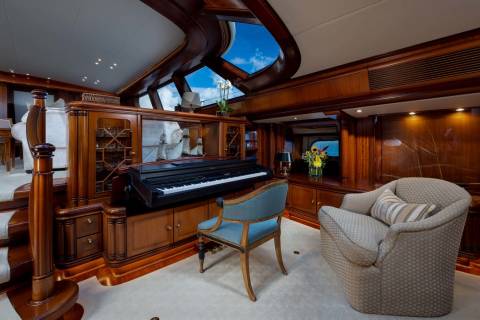 A grand piano sits in the foreground of a luxurious yacht's interior, with a bright blue sky visible through the curved windows in the background.