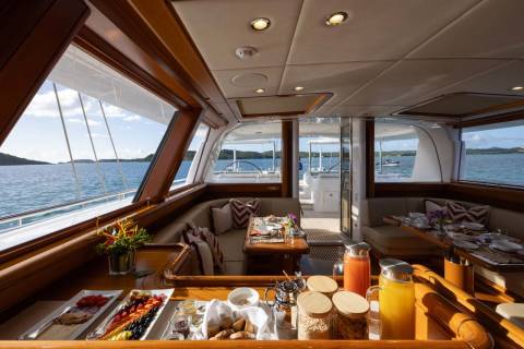 A lavish breakfast spread on a yacht's deck overlooks a calm, blue ocean dotted with islands.