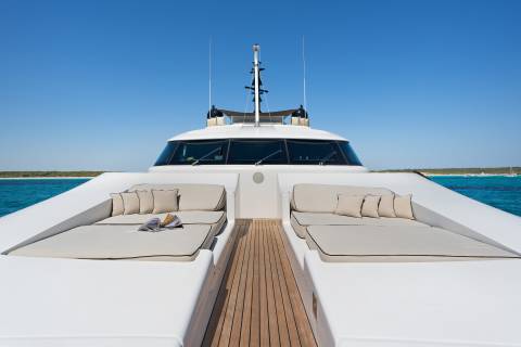 Two lounge areas with beige cushions and pillows sit on the white deck of a yacht, with a clear blue sky and ocean in the background.