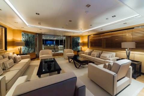 A luxurious living area features cream-colored sofas and armchairs surrounding black coffee tables in the foreground, with wooden blinds and decorative wall panels in the background.