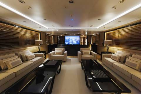 Two long white sofas and black coffee tables face each other in the foreground of a luxurious lounge, with a large screen displaying a city skyline in the background.