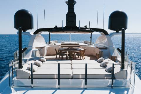 A luxury yacht deck features white lounge seating and a wooden dining area in the foreground, set against a vast blue ocean and clear sky in the background.