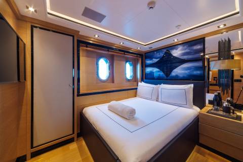 A neatly made bed with a rolled towel sits in the foreground of a luxury yacht cabin, featuring a large ocean-themed artwork and portholes in the background.