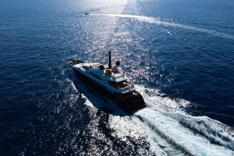 A sleek black and white yacht cuts through shimmering blue water, leaving a white wake behind it while a small boat trails in the distance.