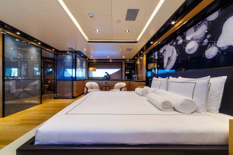 A large bed with white linens occupies the foreground of a modern luxury suite, while the background features a seating area and sleek glass partitions.
