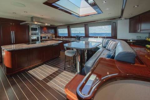 A luxurious yacht interior features a polished wood kitchen and dining area in the foreground, with a panoramic view of the ocean through large windows in the background.