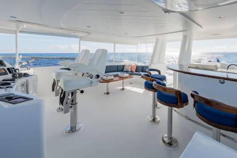 A modern yacht deck features white captain's chairs and a bar area in the foreground, overlooking a calm blue ocean under a bright sky in the background.