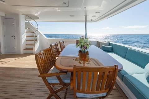A wooden dining table and chairs sit on a yacht deck in the foreground, with a blue cushioned lounge and the open ocean in the background.
