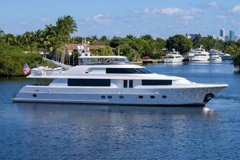 A sleek white yacht floats on a calm blue waterway, with lush tropical greenery and a distant city skyline under a bright sky.