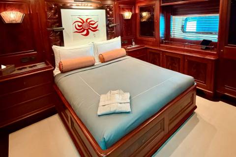 A bed with light blue linens and orange bolster pillows sits in a luxurious wood-paneled room featuring a red sun artwork and a window in the background.