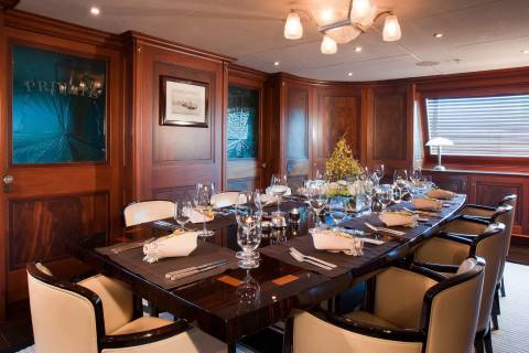A formal dining table set for a meal sits in the foreground of a luxurious room with polished wood paneling and a window in the background.
