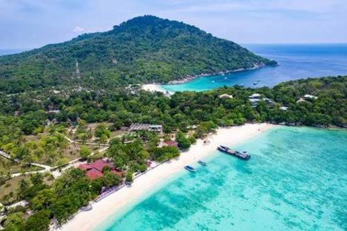 A pristine white sand beach with turquoise waters and boats in the foreground leads to a lush, green, mountainous island in the background.