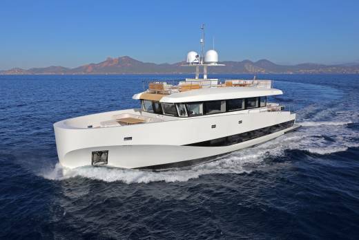 A sleek white yacht cuts through the deep blue sea with a mountainous coastline in the distance.