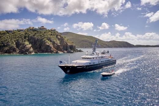 A luxurious yacht cruises through sparkling blue waters near a verdant, cliffside estate under a bright, cloudy sky.