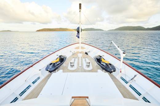Two jet skis are secured on the bow of a luxurious yacht sailing through calm blue waters near a tropical island.