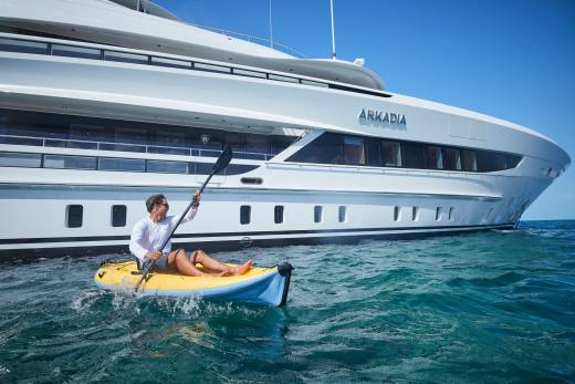 A man paddles a kayak in the clear blue ocean next to the luxurious yacht Arkadia.