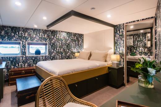 A luxurious cabin aboard a riverboat features bold floral wallpaper, a comfortable bed, and stunning views through porthole windows.