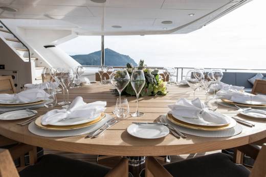 A beautifully set outdoor dining table on a yacht awaits guests with a stunning ocean and mountain backdrop.