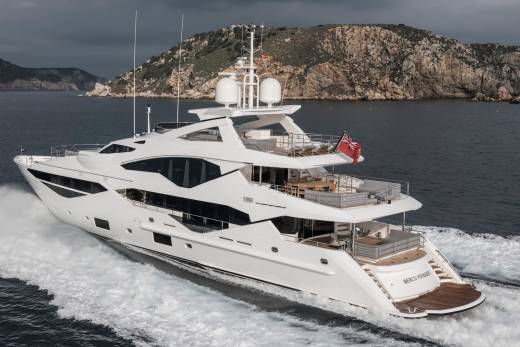 A luxurious white yacht cruises through dark, choppy waters near a rocky, mountainous coastline under a cloudy sky.