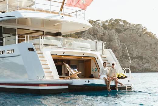 Two women relax on the stern of a luxurious yacht anchored near a rocky, tree-covered coastline.