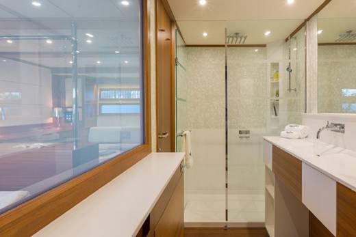 A modern yacht bathroom features a spacious shower, sleek vanity, and a glass partition overlooking a bedroom.