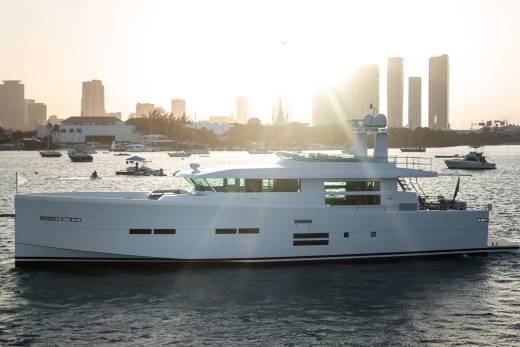 A sleek white yacht cruises through sun-drenched waters with a city skyline in the background.