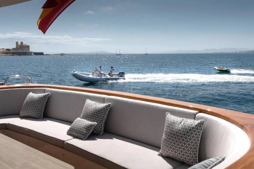 A Spanish flag flutters above a luxury yacht's seating area as a speedboat and a jet ski zip across the sparkling blue ocean.