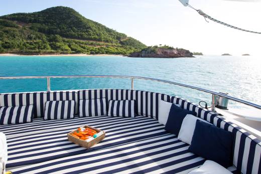 Relax on the striped deck of a yacht with a basket of sunscreen, overlooking a lush green island and turquoise waters.