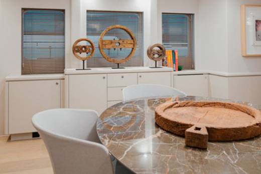 A modern dining area features a marble-topped table with a rustic wooden centerpiece, surrounded by sleek chairs and flanked by decorative ship's wheels.