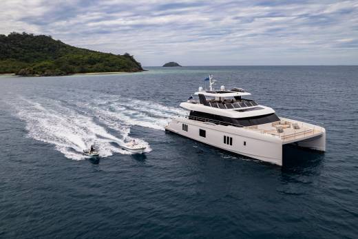A luxurious catamaran yacht cruises through the ocean, trailed by a jet ski and a small tender near a lush, tropical island.