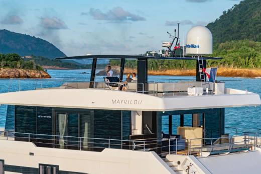 A sleek yacht named MAYRILOU cruises through calm blue waters near a lush, green coastline.