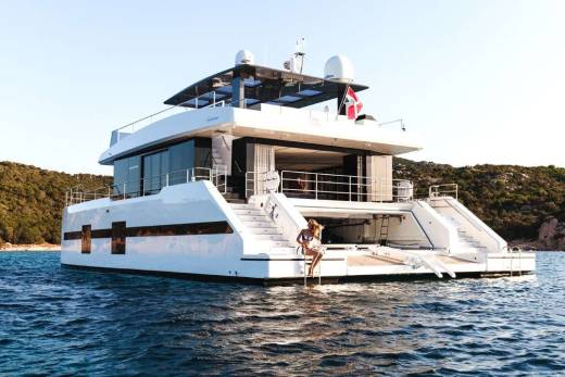 A woman in a bikini descends a ladder from a luxurious yacht anchored near a lush, rocky coastline.