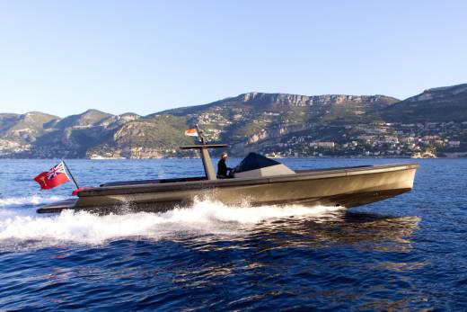 A man pilots a sleek, dark boat through the sparkling blue waters with a mountainous coastline in the background.
