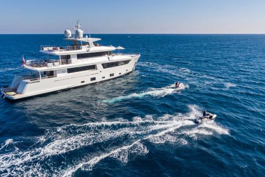 Two jet skis race across the vibrant blue ocean waters, leaving white wakes behind a luxurious white yacht.