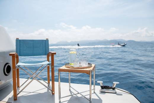 A refreshing pitcher of lemonade and glasses await on a small table beside a director's chair on a boat, with water skiers in the distance.