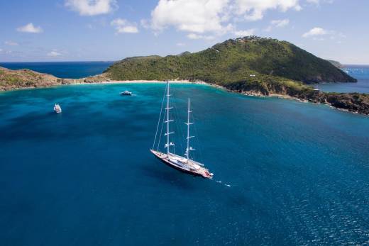 A majestic sailing yacht glides through the crystal-clear turquoise waters off a lush, green island.