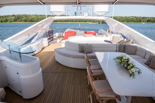 A luxurious yacht deck offers a serene setting with plush seating, a dining table, and a beautiful floral arrangement against a backdrop of blue water and lush green trees.