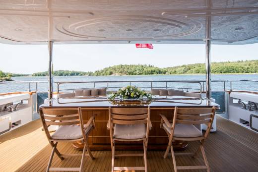 A beautifully set dining table awaits guests on the deck of a luxury yacht, overlooking a serene body of water and lush green shores.