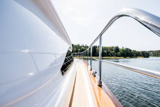 The gleaming white hull of a luxury yacht cuts through calm waters, bordered by a polished railing and lush green coastline.