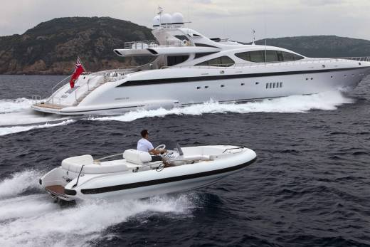A sleek white yacht cruises through choppy dark waters, trailed by a smaller inflatable boat carrying a single person at the helm.