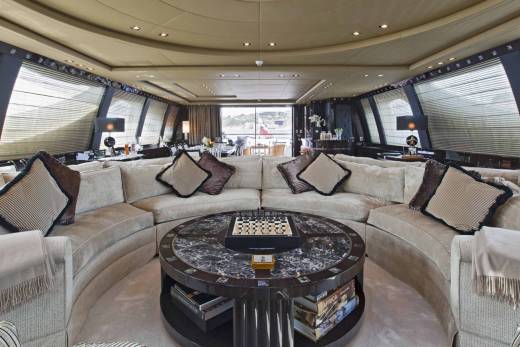 A luxurious yacht lounge features a circular sofa, marble-topped coffee table with a chessboard, and panoramic windows overlooking a scenic landscape.