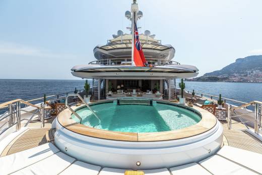 A luxurious yacht features a stunning circular pool on its deck, overlooking the sparkling blue ocean and a distant, picturesque coastline.