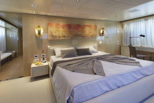 A luxurious bedroom aboard a yacht features a large bed, abstract art, and a desk area with a view of the ocean.