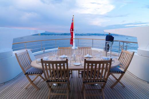 A set dining table awaits on a yacht's deck, overlooking a serene coastal landscape at dusk.