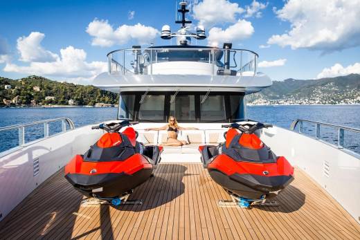 A woman relaxes on a luxury yacht's deck between two vibrant red jet skis, with a picturesque coastal town in the background.