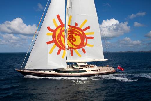 A majestic yacht with a vibrant sun emblazoned on its sail cuts through the deep blue ocean under a bright, cloudy sky.