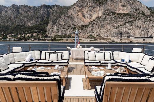 Luxurious white and navy upholstered seating surrounds a wooden table set for tea on a yacht with a dramatic mountain backdrop.