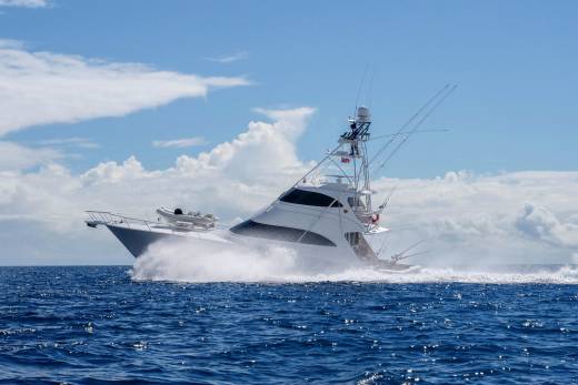A powerful sportfishing yacht cuts through the deep blue ocean, creating a dramatic spray of water under a cloudy sky.