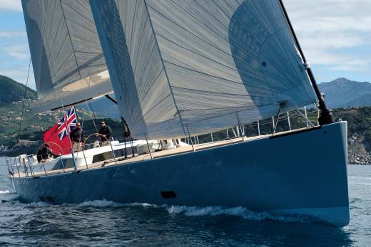 A sleek blue sailboat glides through choppy waters with a Union Jack flag flying proudly against a backdrop of lush green hills.
